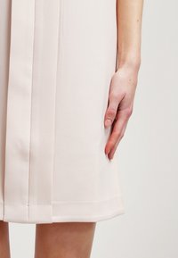 Vaaleanpunainen mekko, joka on valmistettu tekstuuripintaisesta kankaasta, ja siinä on suora leikkaus sekä poimutusdetaljit. Käsivarsi on asetettu sivulle helman lähelle.