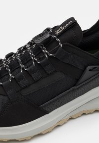 Scarpa sportiva nera con upper testurizzato, lacci elastici, un sistema di chiusura sicuro e una suola in gomma robusta con una suola esterna di un colore più chiaro.