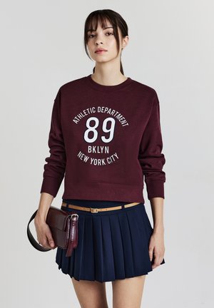 Giovane donna che indossa una felpa bordeaux con la scritta "Athletic Department 89 BKLYN New York City", una gonna blu a pieghe, una cintura beige e tiene una borsa bordeaux.