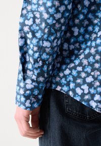 Chemise à manches longues bleue avec un motif floral, présentant des fleurs bleu clair et blanches, des poignets boutonnés et une texture de tissu lisse.