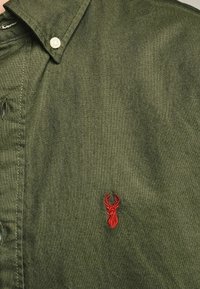 Camicia verde a maniche lunghe in tessuto strutturato, con un ricamo di un cervo rosso sull'area sinistra del petto.