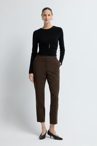 Vrouw staat gekleed in een zwart langslievend ruched top met een sluiting, bruine, op maat gemaakte broek en zwarte kittenheels tegen een effen achtergrond.