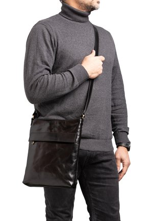 Borsa a tracolla in pelle nera con una superficie liscia, chiusura a pattina e tracolla regolabile, indossata da una persona in un dolcevita grigio.