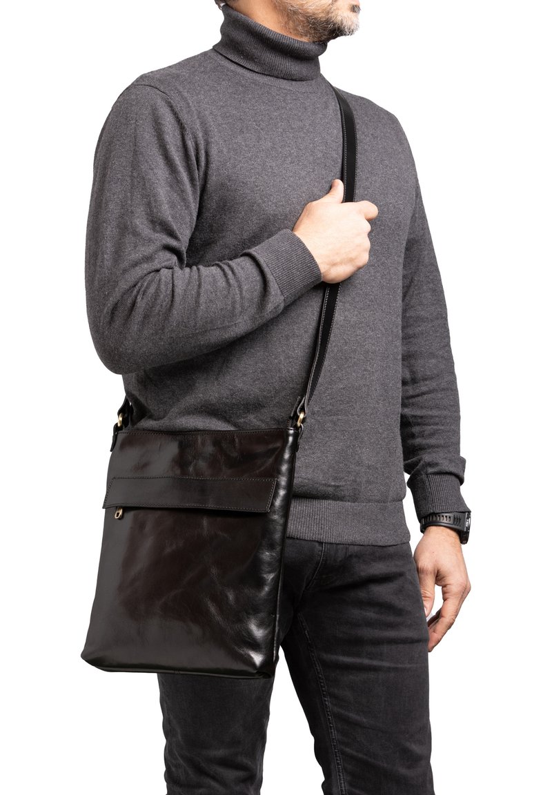 Borsa a tracolla in pelle nera con una superficie liscia, chiusura a pattina e tracolla regolabile, indossata da una persona in un dolcevita grigio.