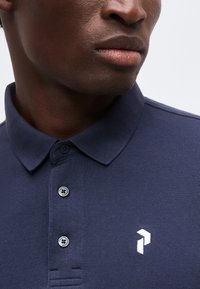 Gros plan d’un homme portant un polo bleu marine avec un col plié, deux boutons et un petit logo géométrique blanc.