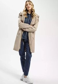 Beżowa wodoodporna kurtka z troczkami, nałożona na dżinsową koszulę, w zestawie z ciemnymi dżinsami i białymi sneakersami, na neutralnym tle.