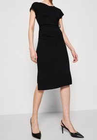 Černé upnuté šaty s krátkými rukávy, hladká textura, délka po lýtka, boční rozparek; v kombinaci s černými lodičkami s došpičatěnými špičkami.