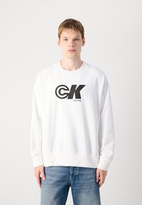 Felpa bianca realizzata in misto cotone, con un logo circolare nero e il testo "Calvin Klein" sul davanti. Vestibilità casual e polsini a costine.