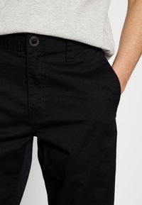 Black cotton chinos with a smooth texture, featuring a front button and zipper, and side pockets. The fabric is medium weight.