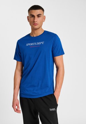 Mann trägt blaues T-Shirt mit der Aufschrift "SPORTS DEPT." und schwarze Hummel-Hose, steht vor einem schlichten hellen Hintergrund.