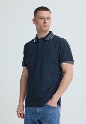 Man met kort haar die een navyblauw poloshirt draagt met gestreepte kraag en manchetten, en blauwe spijkerbroek, staand met één hand in de zak.