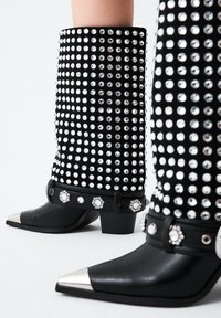 Botas negras hasta la rodilla con tachuelas plateadas y rhinestones, con una punta metálica puntiaguda, sobre un fondo blanco.