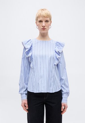 Kvinde med kort blondt hår iført blå og hvidstribet bluse med flæser ved skuldrene og sorte bukser, stående mod en ensfarvet lys baggrund.