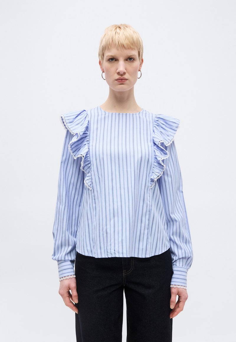 Γυναίκα με κοντά ξανθά μαλλιά, φορώντας μπλούζα μπλε και λευκές ρίγες με βολάν στους ώμους και μαύρο παντελόνι, στέκεται μπροστά σε απλό φωτεινό φόντο.