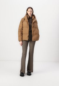 Giacca bomber marrone con cappuccio, caratterizzata da un design trapuntato e chiusure a pressione. Abbinata a pantaloni a campana con motivo pied de poule e scarpe nere.