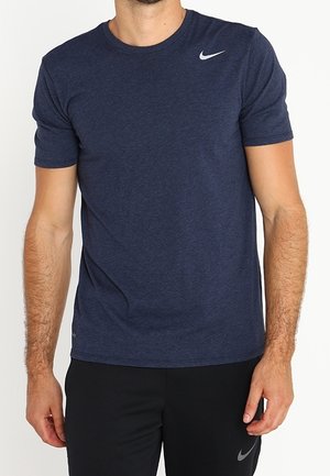 Mann trägt ein dunkelblaues Nike-T-Shirt mit kurzen Ärmeln und schwarze Nike-Hose mit sichtbaren Logos, steht vor einem schlichten Hintergrund.