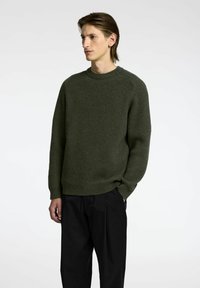 Suéter de punto verde con canalé, cuello redondo y mangas largas, combinado con pantalones negros, mostrado sobre un fondo simple.