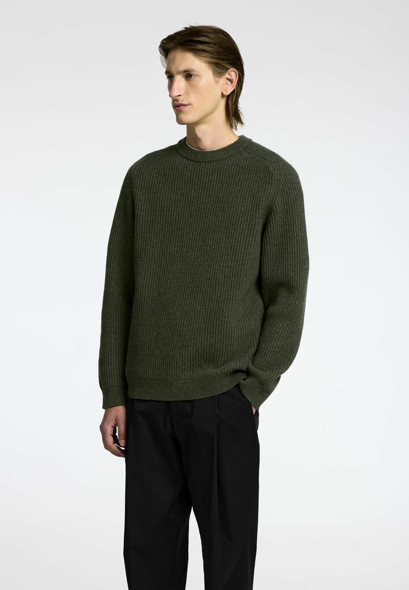 Suéter de punto verde con canalé, cuello redondo y mangas largas, combinado con pantalones negros, mostrado sobre un fondo simple.