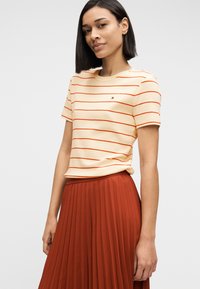 Γυναίκα με κρεμ μπλουζάκι με λεπτές κόκκινες οριζόντιες ρίγες και σκουρόχρωμη πλισέ φούστα, που στέκεται μπροστά σε απλό ανοιχτό φόντο.