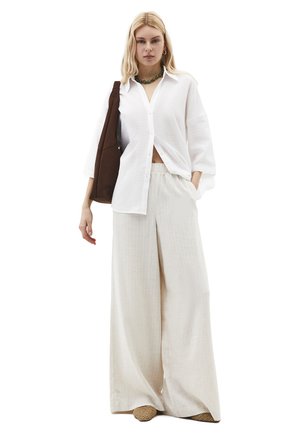 Mujer rubia con camisa blanca de botones holgada, pantalones anchos color crema, collar verde de cuentas, bolso marrón de hombro y zapatos tejidos.