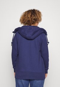 Marineblauwe hoodie met trekkoord, zachte textuur, ontspannen pasvorm en geribbelde onderkant. Achteraan is er een eenvoudig ontwerp zonder logo's.