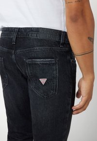 Juodi džinsai su šiek tiek nusidėvėjusiu tekstūru, turintys GUESS logotipo tatuiruotę ant galinio kišenės ir iškilnų juosmens dirželį.