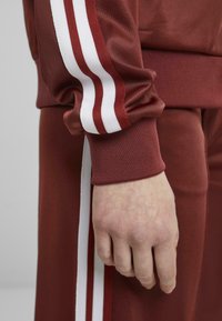 Maroon tracksuit with white and red stripes, showcasing a textured fabric and fitted cuffs. Hand resting against the fabric, showing detail.