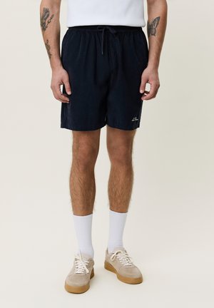 Mann iført svarte shorts med snor, hvite sokker, beige joggesko og hvit skjorte, viser tatoveringer på begge underarmene.