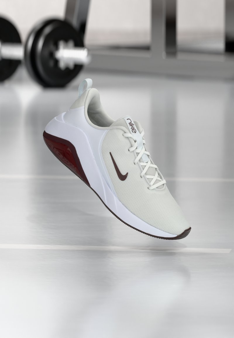 Sapatilha branca desportiva com parte superior em malha texturizada e detalhes em bordô. Apresenta uma sola branca em contraste e um logo da Nike em destaque.