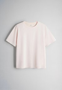 Plain light pink short-sleeve cotton t-shirt with round neckline hanging against a light gray wall.