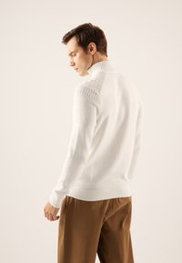 Suéter de punto blanco con cuello alto, que presenta detalles texturizados en los hombros y puños acanalados, combinado con pantalones marrones.