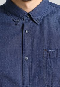 Mörkblå knapp-up skjorta i ett strukturerat tyg, med en klassisk krage, två knappar och en bröstficka med subtil märkning.