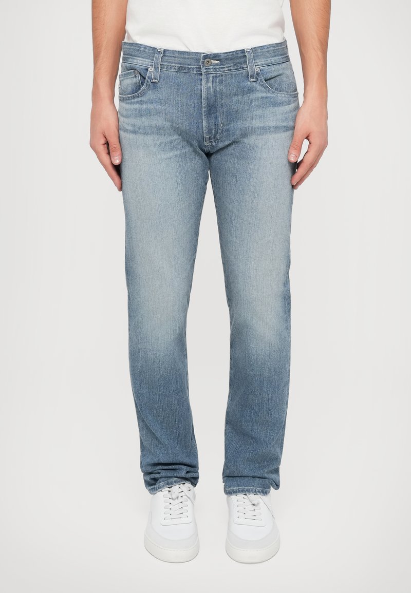 Ανοιχτό μπλε τζιν παντελόνι με ίσια γραμμή, πέντε τσέπες, διακριτική φθορά και κανονικό κλείσιμο με κουμπί και φερμουάρ, τα οποία είναι συνδυασμένα με λευκά αθλητικά παπούτσια.