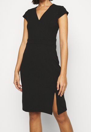 Femme portant une robe noire ajustée avec des manches courtes, un décolleté en V et une fente sur la cuisse droite.