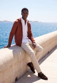 Homme portant des lunettes de soleil, un sweat à capuche marron, une chemise blanche et un pantalon beige, assis sur une rambarde en pierre au bord d'une mer ensoleillée avec des collines lointaines en arrière-plan.