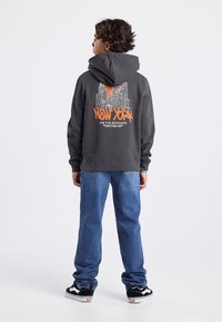 Sudadera gris con un gráfico de rascacielos de Nueva York en negro y naranja, combinada con vaqueros rectos azules y zapatillas negras.