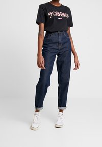 Camiseta de algodón negra con el logo "TOMMY" en rojo y blanco, combinada con jeans de mezclilla oscuros de cintura alta, con puños enrollados, y zapatillas blancas.