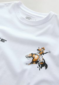 Narancssárga-fekete cowboy lovaglás közben egy sprőd lovon, fehér pólón, a nyakrésznél, részlegesen látható szöveggel.