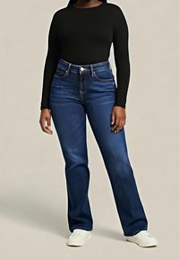 Personne portant un jean bleu foncé taille haute, un haut noir à manches longues et des baskets blanches, debout devant un fond uni.