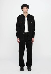 Camisa de algodón negra de manga larga con dos bolsillos en el pecho y botones en tono dorado, combinada con pantalones y zapatos negros a juego. Camiseta blanca visible.
