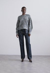 Grå tekstureret sweater med lange ærmer, parret med mørkeblå lige ben jeans og sorte højhælede sko, sat mod en ensfarvet baggrund.