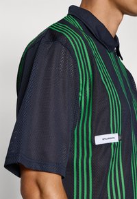 Kortermet marineblå mesh-skjorte med grønne vertikale striper, kragehals og en frontlabel med teksten "STUDENTER."