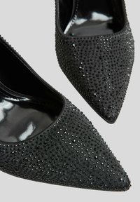 Czarne czółenka na wysokim obcasie z ostrym noskiem, pokryte teksturowaną tkaniną i ozdobione błyszczącymi kryształkami, z połyskującą podszewką wewnętrzną.