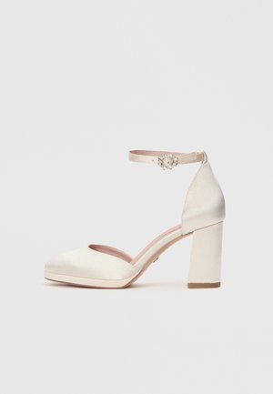 Heel height shoe with a satin finish in off-white. Features a rounded toe and ankle strap with decorative embellishment. Block heel design.