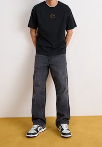 Chemise noire à manches courtes avec col rond et logo doré "Nike Air", associée à un jean gris foncé décontracté et des baskets noir et blanc.