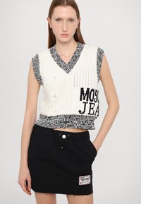 Austā krēmkrāsas krope ar melniem akcentiem, ar V veida kakla izgriezumu, kabelīšu rakstu un "MOSCHINO JEANS" logotipu. Pāris ar melnu mini svārku.