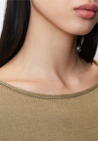 Olijfgroene, gebreide top met een ruime halslijn en een gestructureerde afwerking. Close-up van de kraag en schouderpartij, die de stikdetails benadrukt.