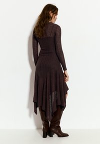 Langærmet, mørkebrun kjole med ribbet tekstur, flydende kanten og asymmetrisk snit, parret med brune støvler op til knæene.
