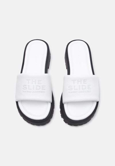 Λευκά σανδάλια με φαρδιές λουρίδες και μαύρες σόλες, με ανάγλυφο κείμενο "THE SLIDE MARC JACOBS" στις επενδυμένες λουρίδες, εμφανιζόμενα από πάνω.