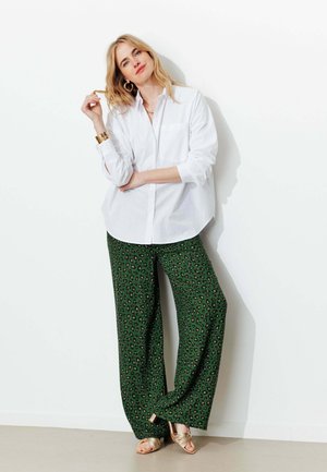 Femme blonde appuyée contre un mur, portant une chemise blanche boutonnée, un pantalon large vert à imprimé léopard, des sandales dorées et des boucles d’oreilles créoles.